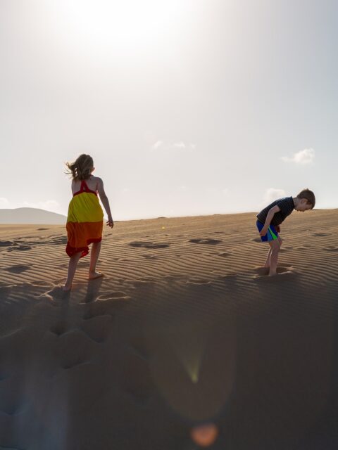 Squeals of kids jumping across the dunes, kicking up clouds of dust with their heels. 
Wind whipping against cheeks, carrying the scent of the sea in shades of choppy turquoise below. 
Whitewashed villages with emerald green painted window panes chipping dry in the searing sun. 
Summer-bleached terracotta roofs. 
Endless slopes rolling across the land.
Volcanic.
Surreal.
Bare. 
A natural sculpture that stretches to the horizon where the ocean pounds against the shore.
Wide-open spaces. 
Baked under the heat.
Stark beauty. 
Silence.
Long stretches of road with no traffic. 
Windmills. 
Cacti. 
Rocks. 
Tufts of dry plants where goats graze. 
Single palm trees stretching high into a brilliant blue sky. 
Each village marked with big white letters that the kids climb on: Betancuria. La Oliva. Antigua. 
This is Fuerteventura. 
The oldest of the Canary Islands. 
Once a place of exile for Spanish write Miguel de Unamuno who described it as “a skeleton of land.”